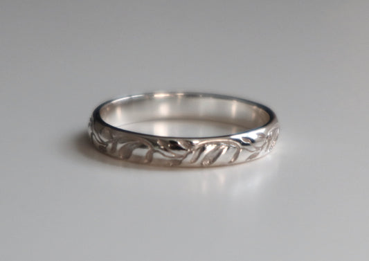 A silver band ring with a textured, vine engraved pattern around the outer edge, shown on a neutral gray background.